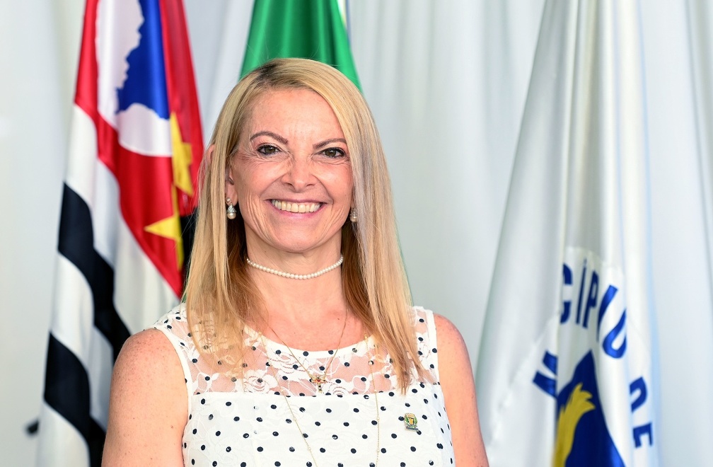 Fotografia da secretária Alexandra Caprioli, de pé, com as bandeiras de Campinas e do Estado de São Paulo ao fundo