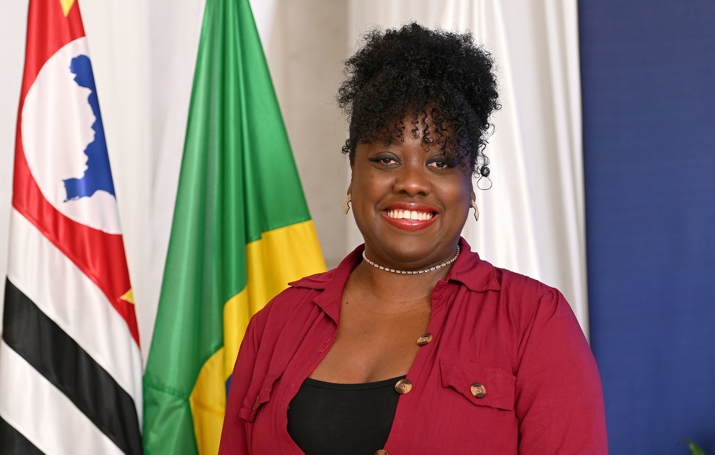 Fotografia da secretária Eliane Jocelaine Pereira, de pé, com braço direito levemente apoiado no encosto de uma cadeira