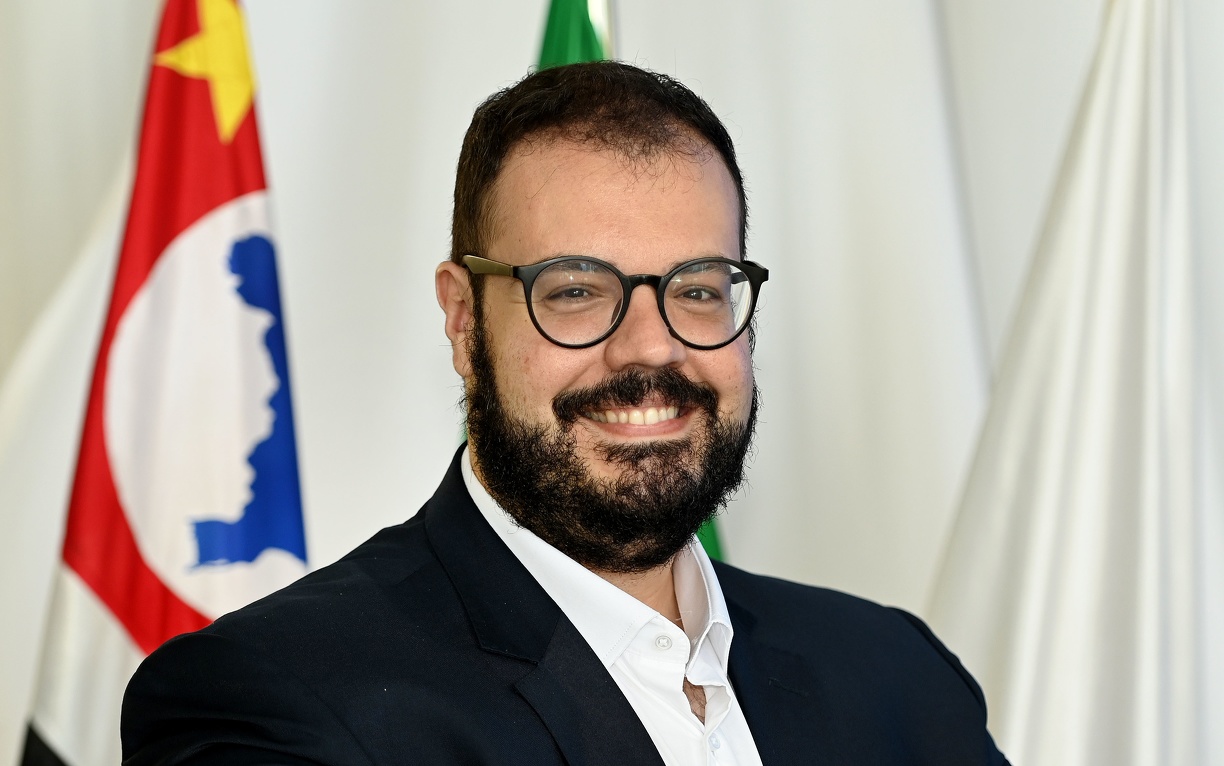 Fotografia do secretário Fernando de Caires, em pé, com as bandeiras de Campinas, do Estado de São Paulo e do Brasil ao fundo