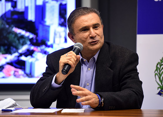 Fotografia do secretário Aderval Fernandes Júnior , sentado, com microfone à mão, com telão exibindo imagens durante uma apresentação feita por ele.