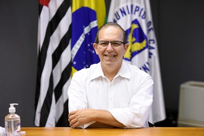 Fotografia do secretário Aurílio Sérgio Costa Caiado, sentado, com as bandeiras de Campinas, do Estado de São Paulo e do Brasil ao fundo