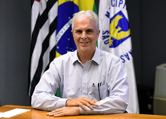 Fotografia do secretário Carlos José Barreiro, sentado, com as bandeiras de Campinas, do Estado de São Paulo e do Brasil ao fundo