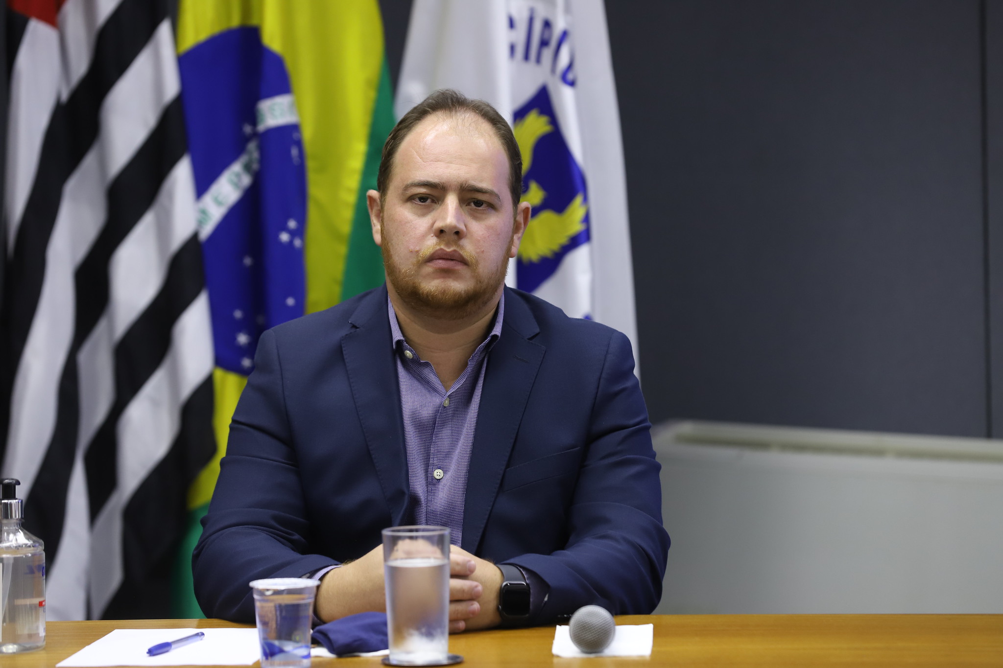 Fotografia do secretário Christiano Biggi Dias, sentado, com as bandeiras de Campinas, do Estado de São Paulo e do Brasil ao fundo