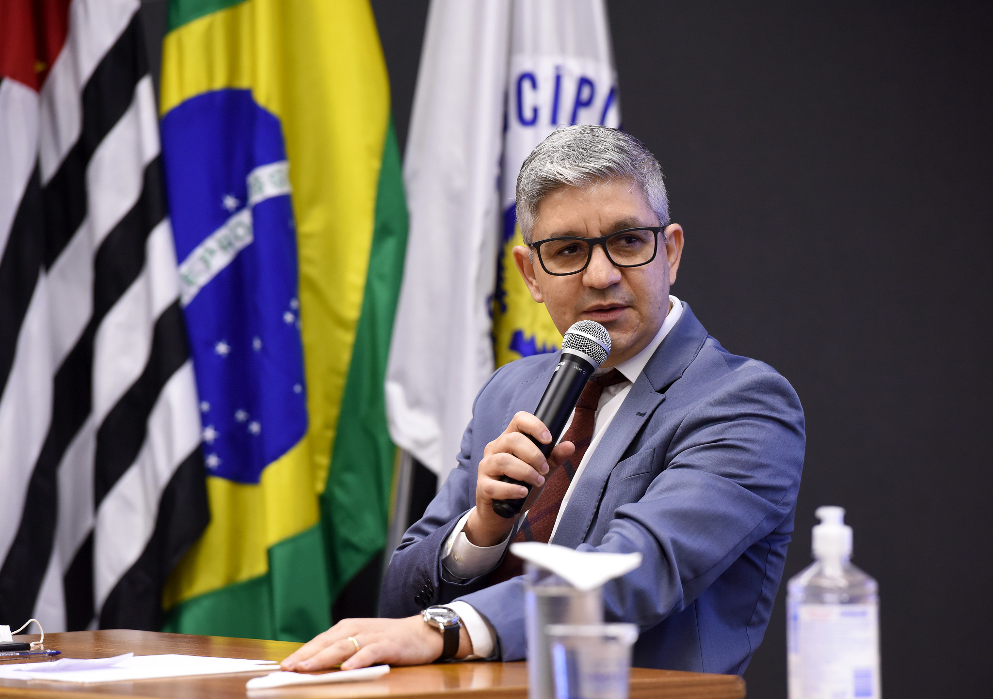 Fotografia do secretário Aurílio Sérgio Costa Caiado, sentado, falando ao microfone, com as bandeiras de Campinas, do Estado de São Paulo e do Brasil ao fundo