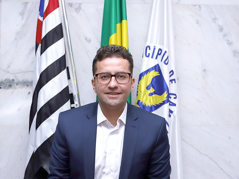 Fotografia do presidente da EMDEC, Vinícius Issa Lima Riveretei, sentado, com as bandeiras de Campinas e do Brasil ao fundo