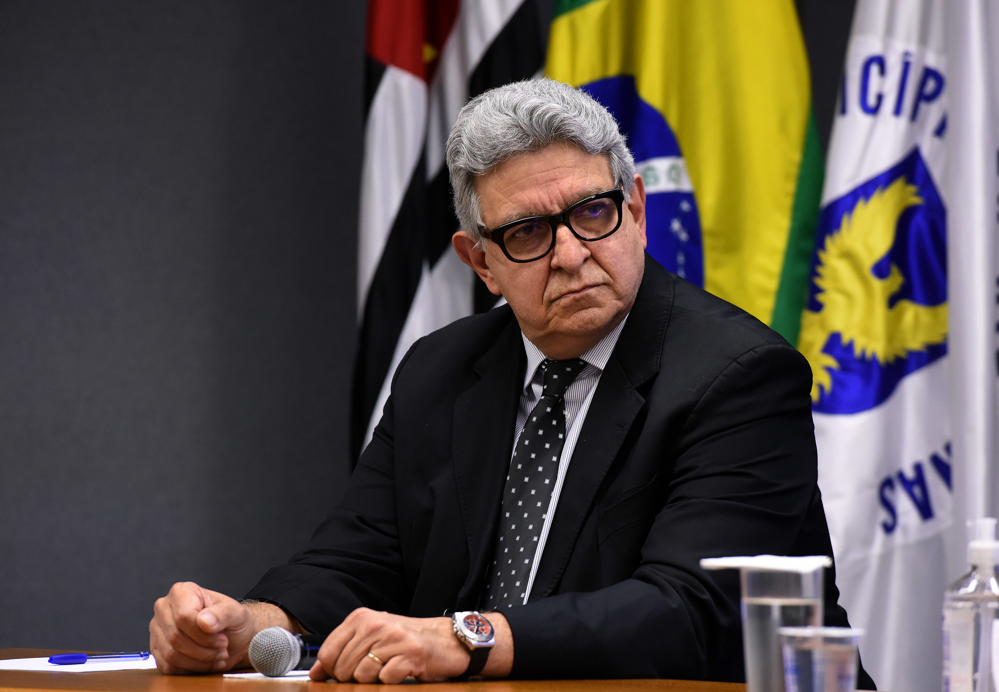 Fotografia do presidente Sérgio Bisogni, sentado, com as bandeiras de Campinas, do Estado de São Paulo e do Brasil ao fundo.