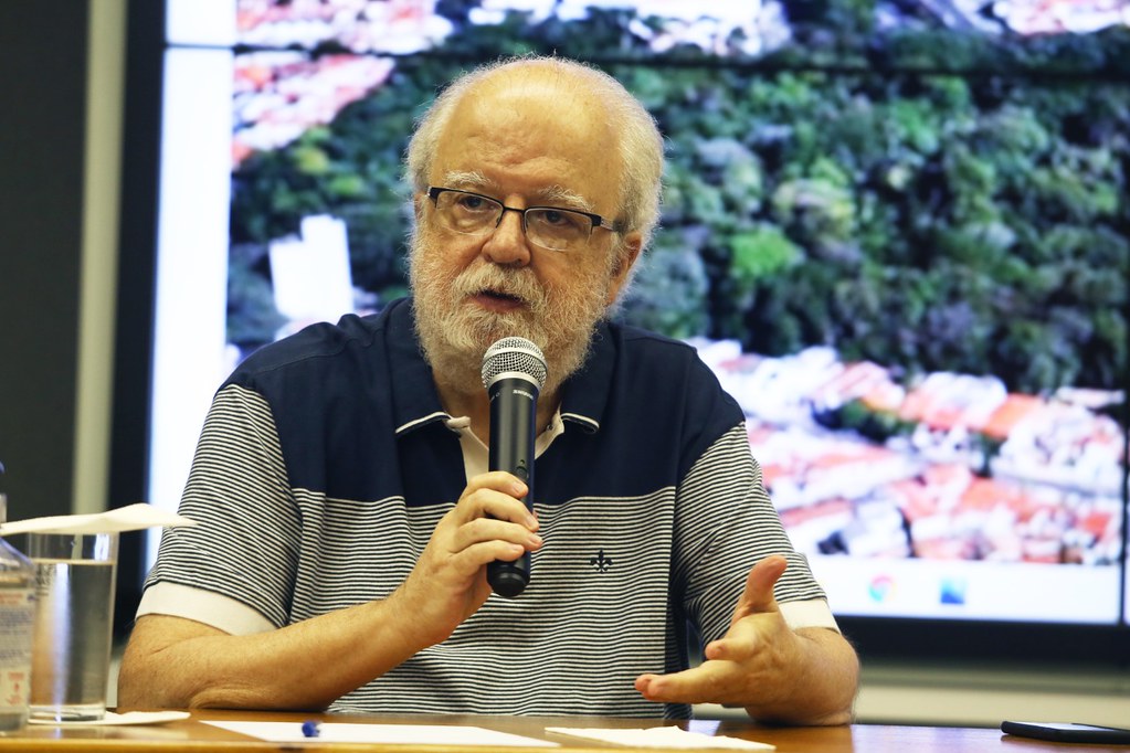 Fotografia do secretário José Tadeu Jorge, sentado, falando ao microfone com uma tela projetando uma apresentação ao fundo.